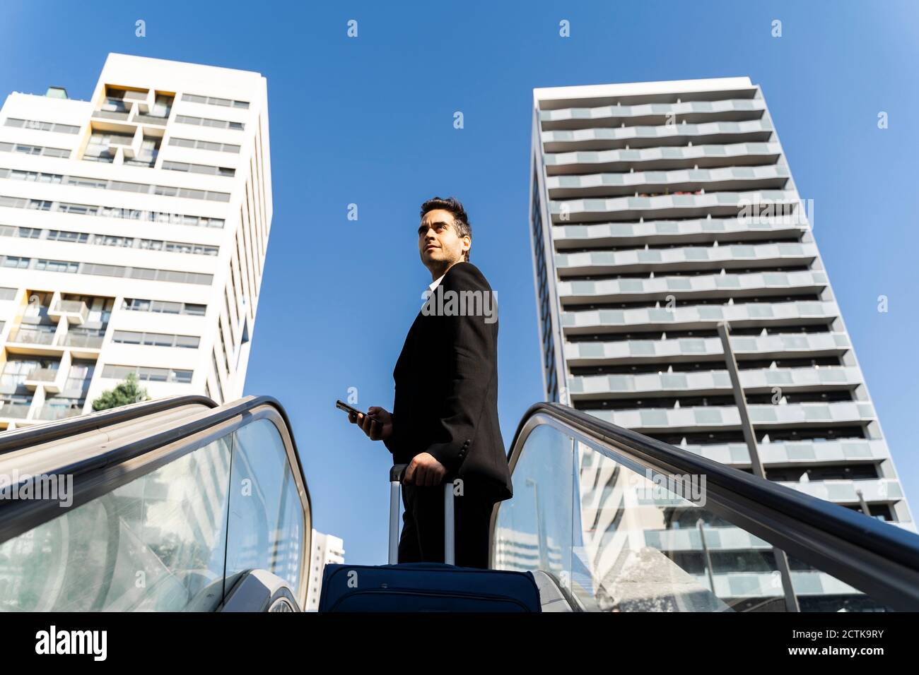 Homme d'affaires avec valise à l'écart en se tenant sur l'escalier roulant contre ciel dégagé en ville Banque D'Images