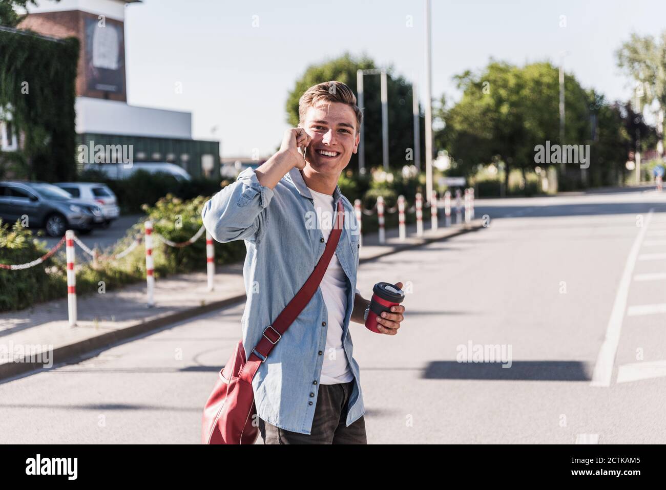 Jeune homme souriant parlant sur téléphone portable en traversant la rue en ville Banque D'Images