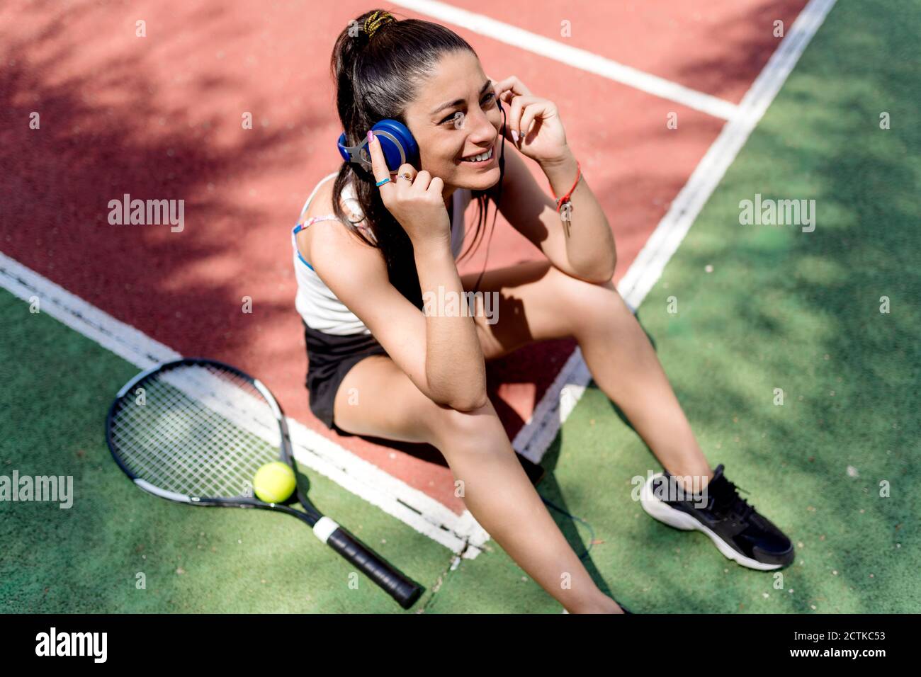 Femme souriante, joueur de tennis, écoutant de la musique tout en étant assise sur le sol en cour Banque D'Images