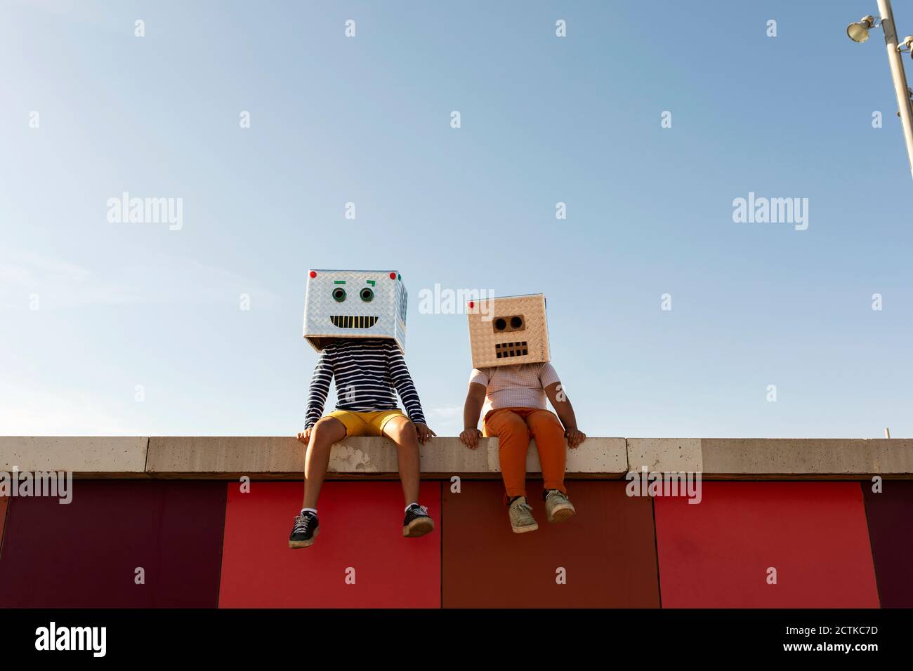 Amis portant des masques robotisés assis sur le mur de retenue contre clair ciel bleu Banque D'Images