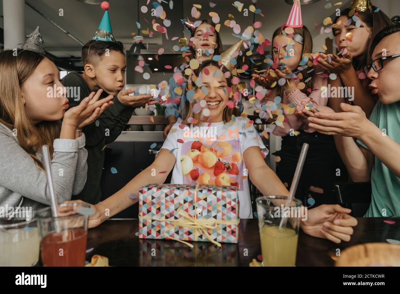 Des amis heureux jetant des confetti sur une adolescente assise avec un anniversaire cadeau à la table à manger Banque D'Images