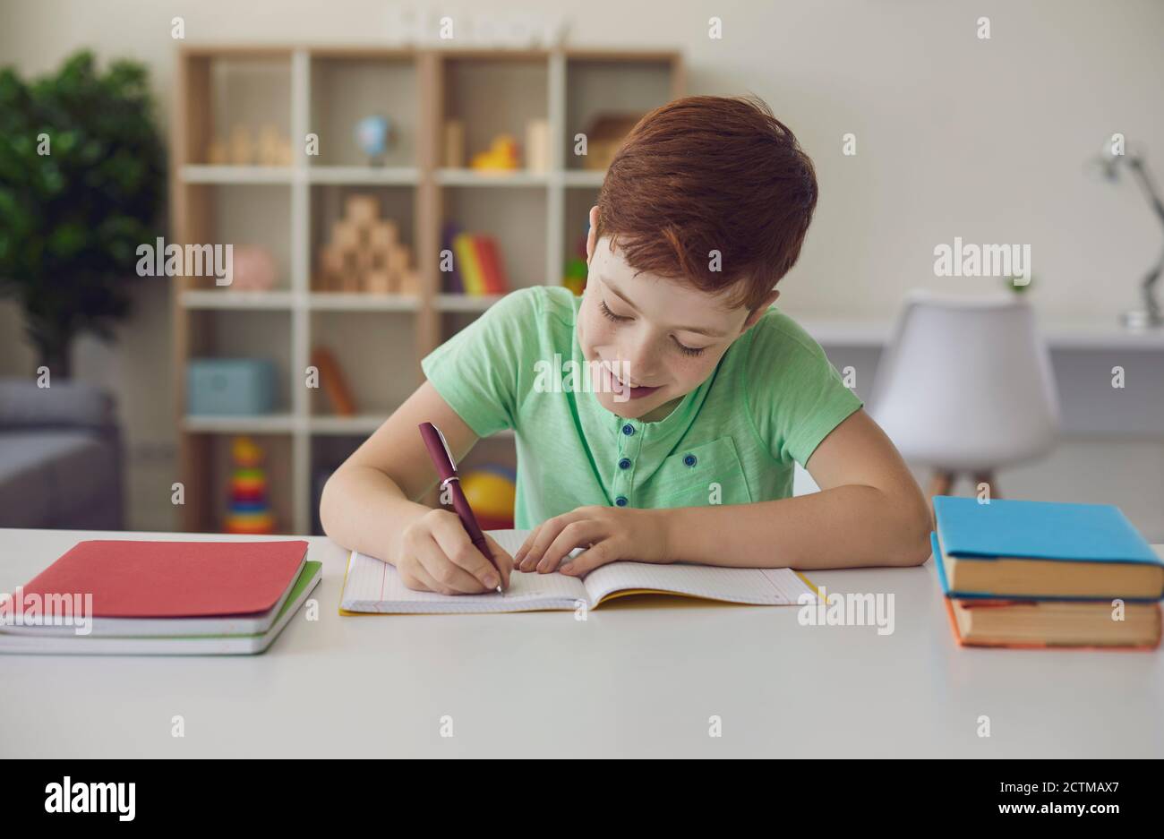 Un petit garçon souriant qui fait ses devoirs assis à un bureau dans sa chambre lumineuse. Banque D'Images
