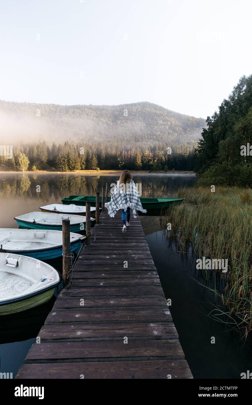Femme debout sur la jetée en bois près du lac avec des bateaux, et appréciant la beauté de la nature au lever du soleil dans la forêt. Banque D'Images