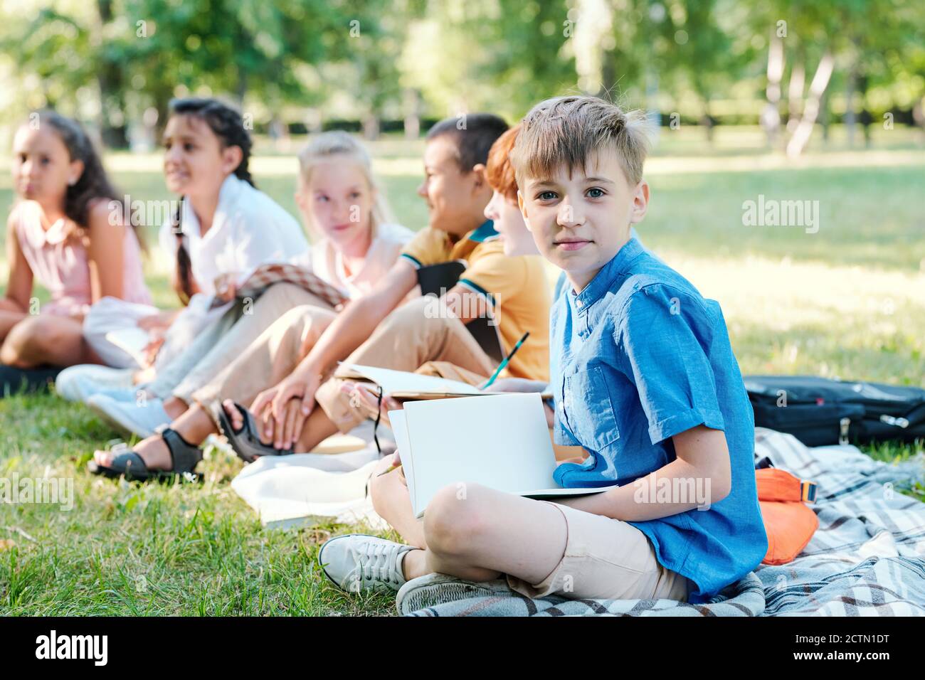 Portrait du contenu garçon en chemise bleue avec croix jambes sur le tissu écossais avec les camarades de classe et tenue du cahier à cours d'école extérieure Banque D'Images