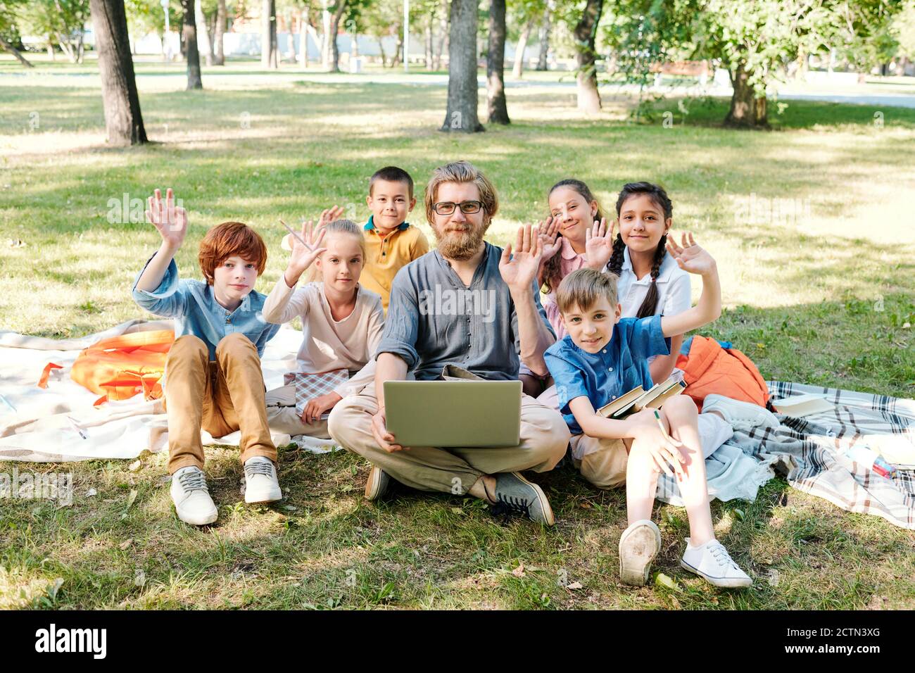 Groupe d'enfants et leur professeur assis sur des couvertures dans le parc et en se faisant passer les mains à la caméra, cours de plein air créatif Banque D'Images