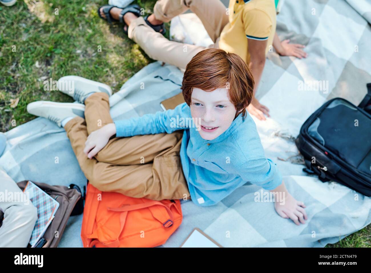 Vue en grand angle d'un écolier souriant à tête rouge dans une chemise bleue assis avec une sacoche sur une couverture et passer du temps avec des amis au pique-nique après l'école Banque D'Images
