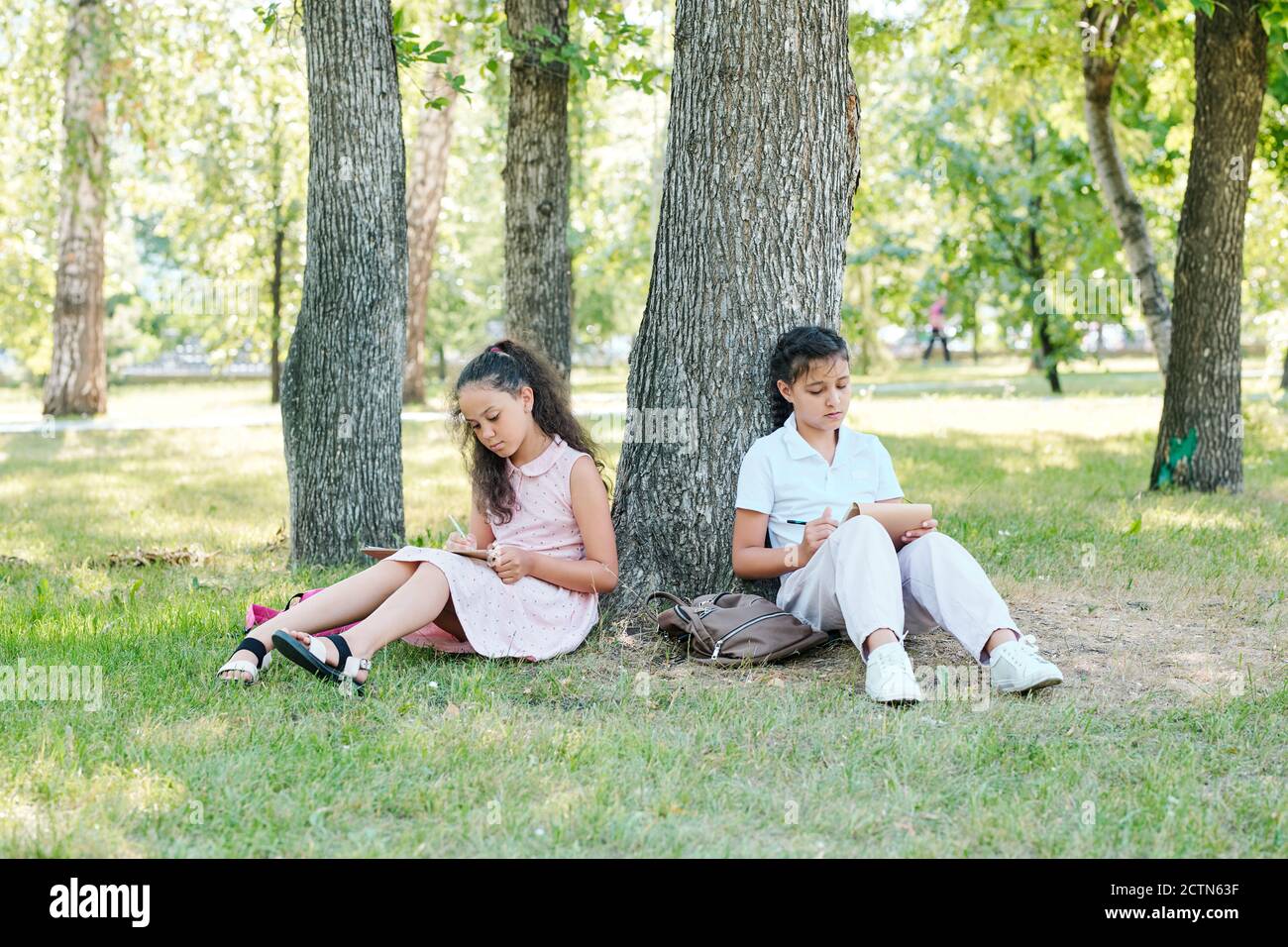 Filles d'école de race mixte focalisées assis à l'arbre dans le parc et exécution de la tâche avant la classe scolaire dans le parc Banque D'Images