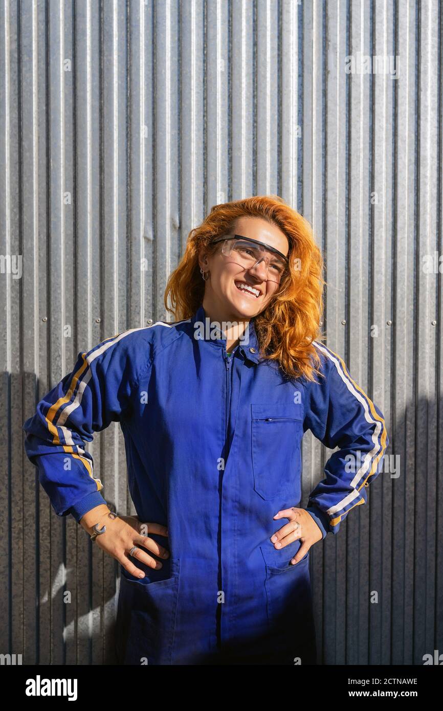 Femme ingénieuse en lunettes de protection et vêtements de travail à proximité mur en métal à l'usine et regarder loin tout en profitant de la pause pendant le travail Banque D'Images