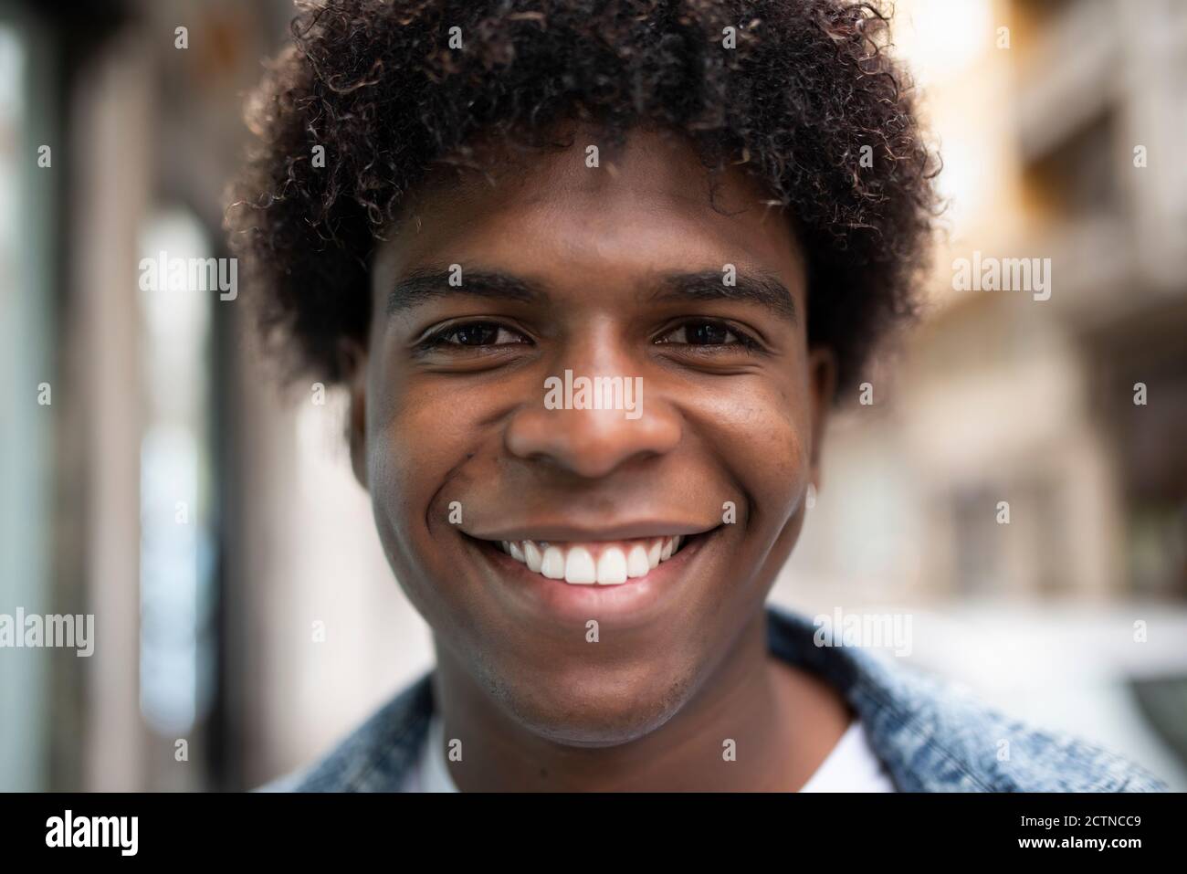 Homme afro-américain positif avec une coiffure afro et un sourire charismatique regarder l'appareil photo tout en se tenant sur un arrière-plan flou de la ville Banque D'Images