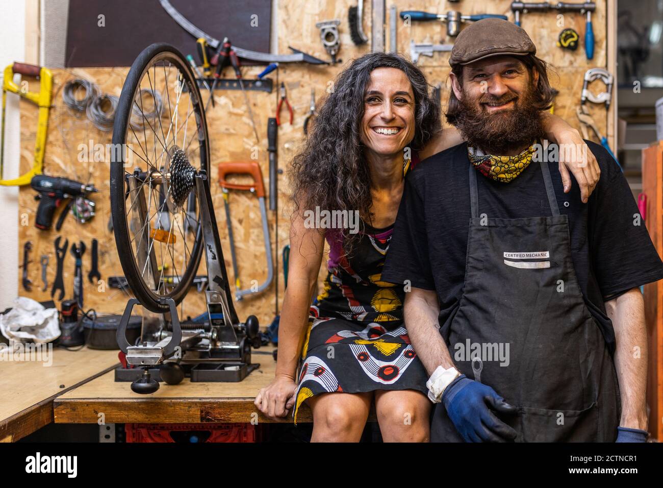 Couple ethnique positif des propriétaires de l'atelier de service de vélo regardant à l'appareil photo et souriant amical contre les intempéries mur avec divers réparer les instruments Banque D'Images