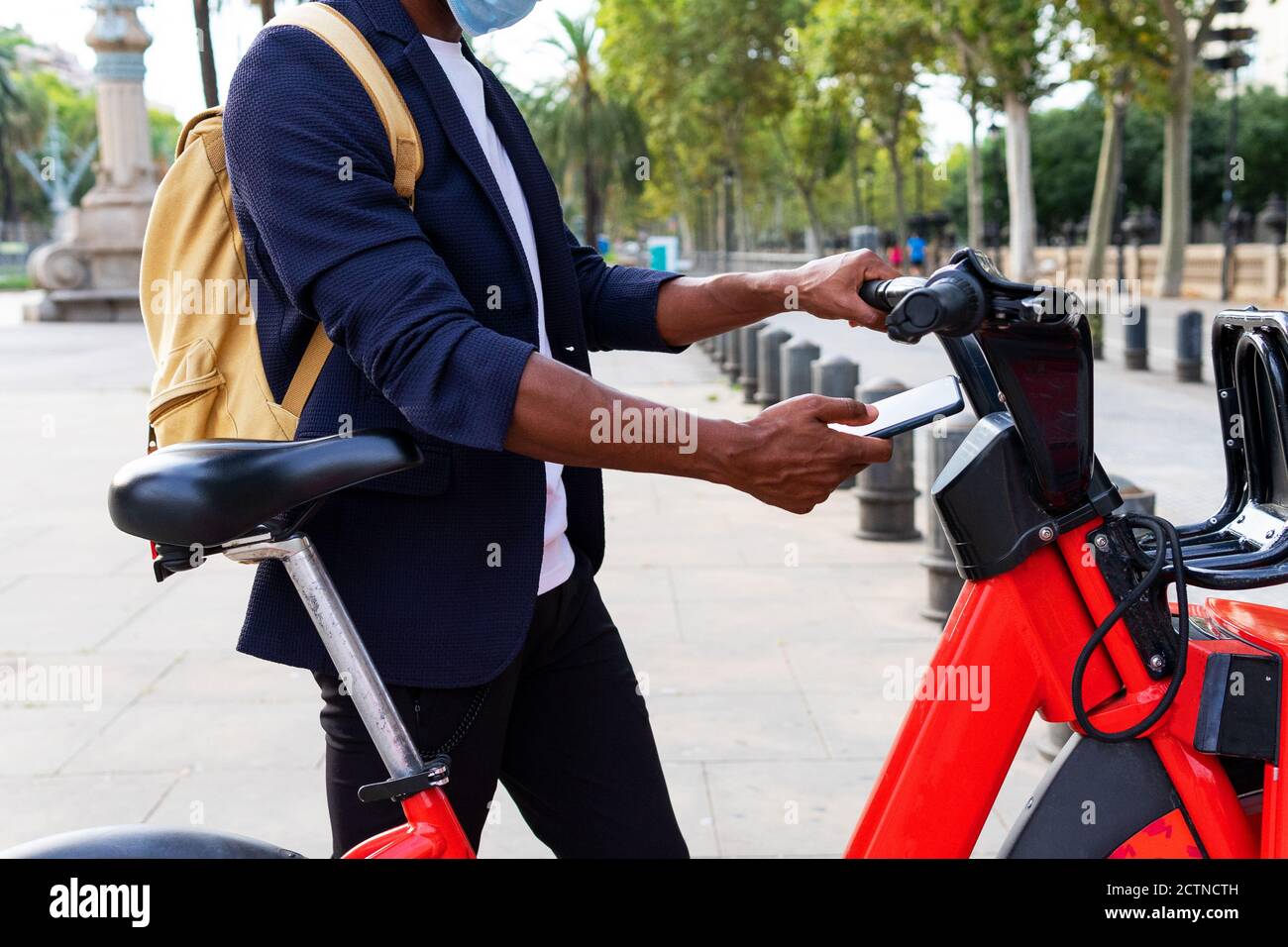 Homme noir en costume et masque en utilisant sa cellule téléphone pour louer un vélo public dans la rue Banque D'Images