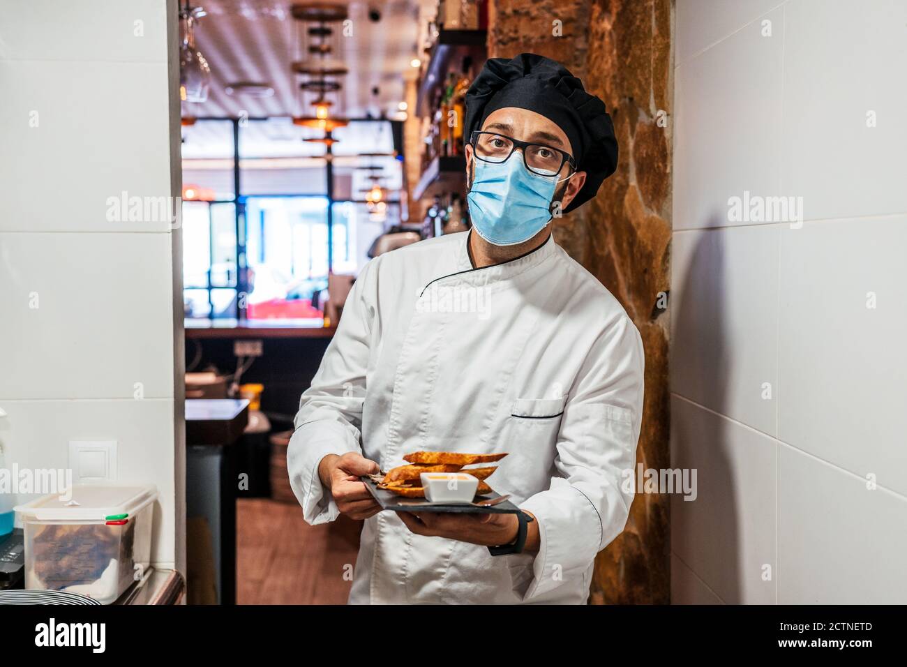 Chef masculin sérieux en uniforme et masque médical debout avec délicieux en-cas sur le plateau en ardoise dans le café et regarder appareil photo Banque D'Images