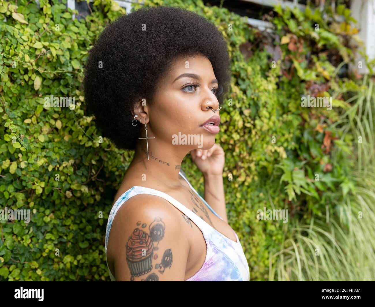 Vue latérale d'une femme afro-américaine élégante avec tatouages et piercing debout dans le parc urbain en été et regarder loin Banque D'Images