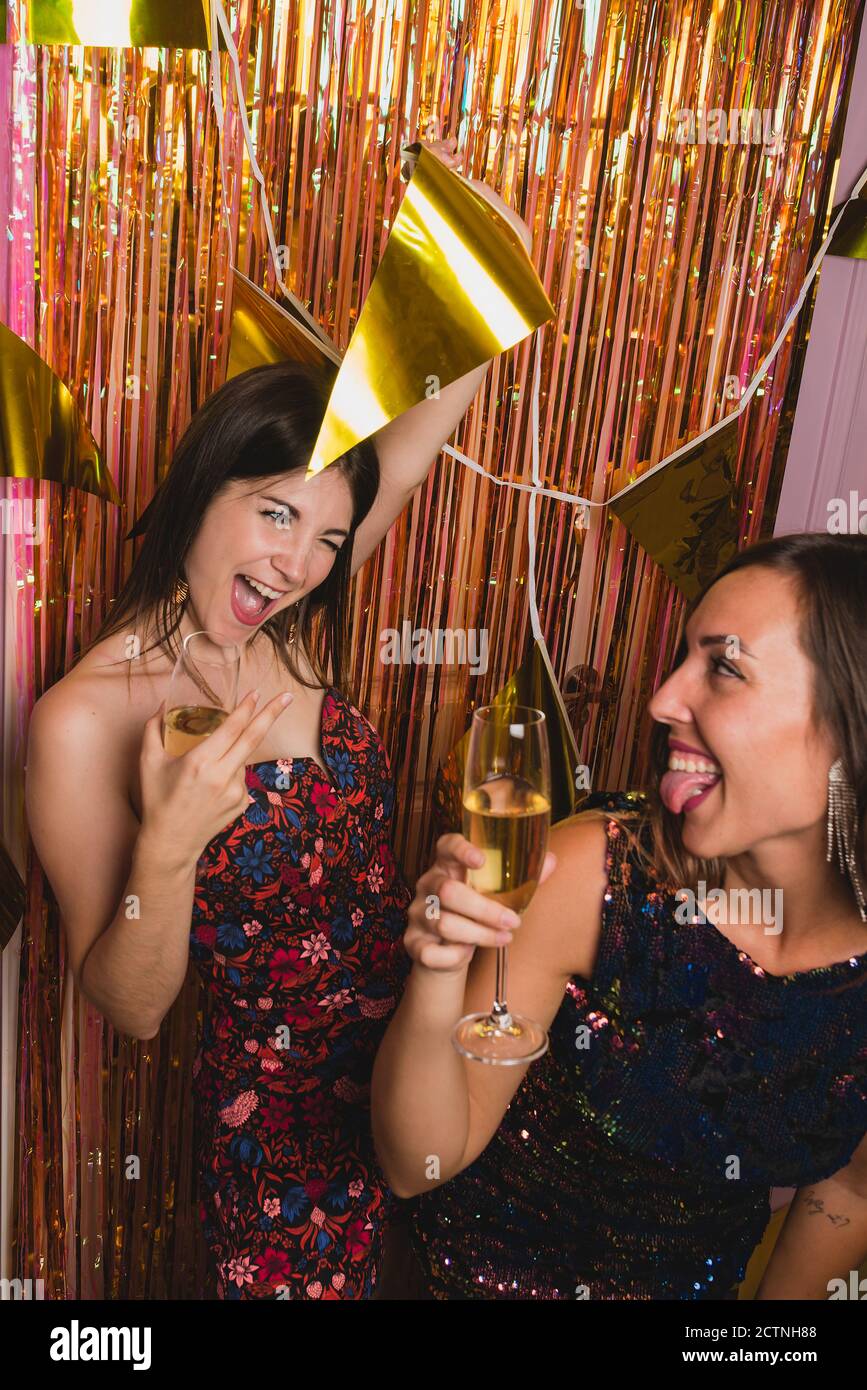 Jeunes amies gaies dans des robes de fête stylées qui applaudissent des verres de champagne et regarder l'un l'autre tout en appréciant Fête du nouvel an Banque D'Images