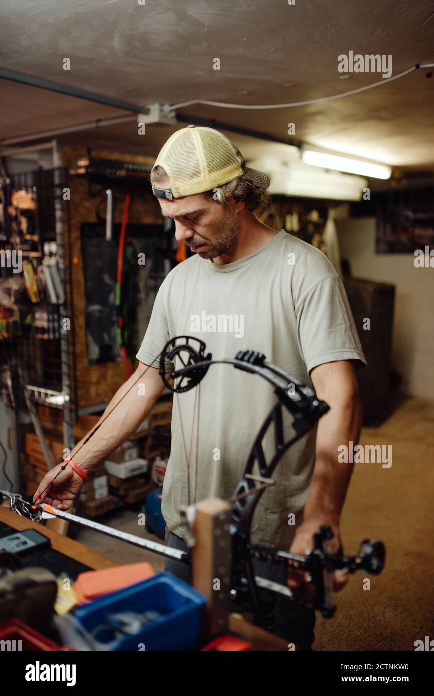 Vue latérale d'un chasseur de sexe masculin plein d'activité en train de régler l'arc composé avec flèche pendant que vous vous tenez dans le garage et que vous vous préparez pour la prise de vue Banque D'Images