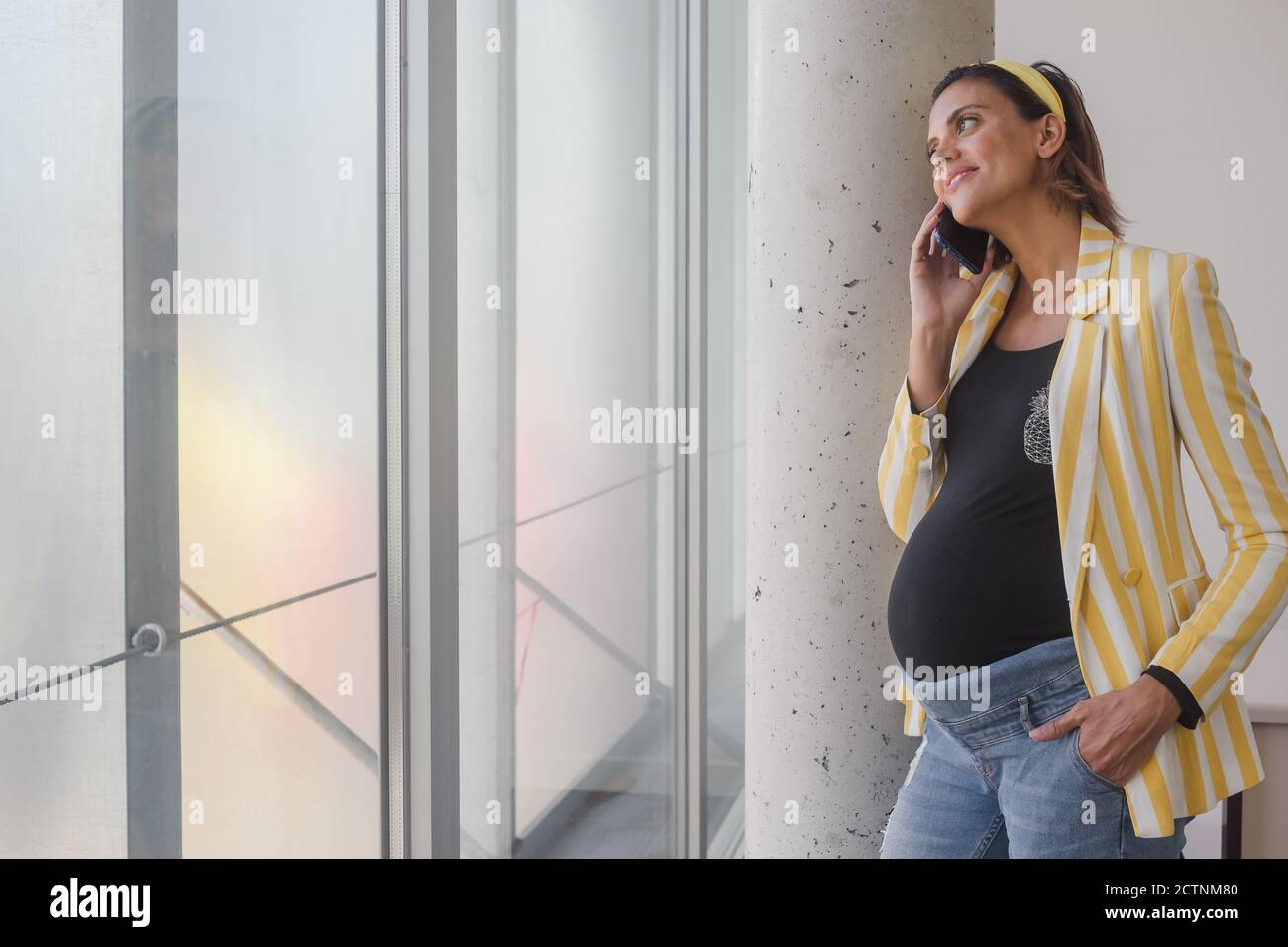 Vue latérale d'une jeune femme enceinte gaie dans une tenue élégante parler sur un téléphone portable et sourire en se tenant près du verre mur dans un bâtiment contemporain Banque D'Images