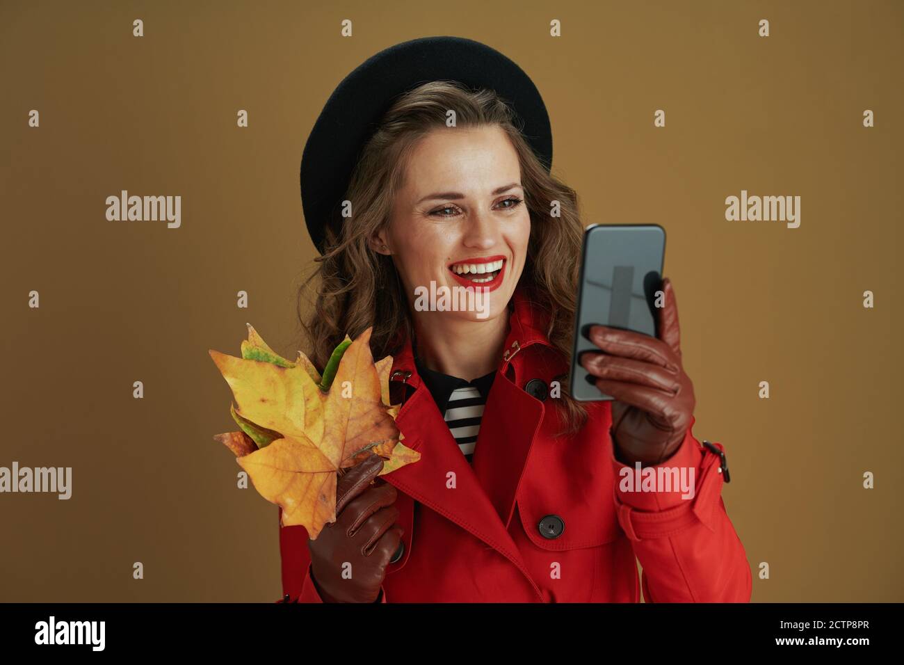 Bonjour octobre. Femme souriante et branchée de 40 ans en manteau rouge et  béret noir avec gants en cuir et feuilles d'érable jaune d'automne à l'aide  de l'appli pour smartphone Photo Stock -