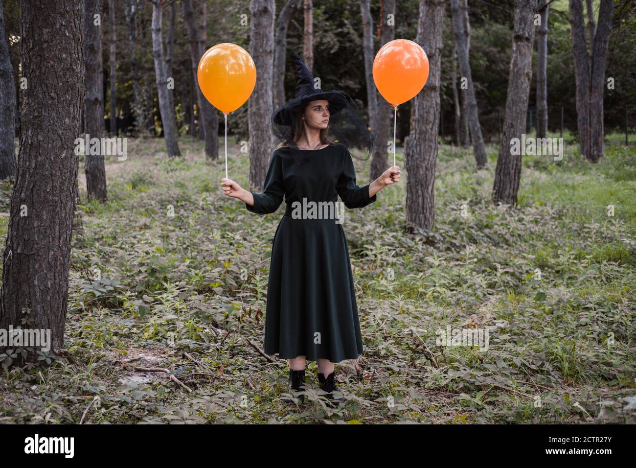 Jeune mignonne belle femme en robe sombre et chapeau de sorcière tient des ballons d'orange dans ses mains. Costume de fête d'Halloween. Forêt, parc avec arbres d'automne Banque D'Images