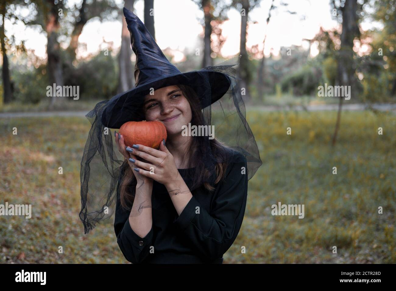 Une jeune femme en robe noire et chapeau de sorcière tient une citrouille dans ses mains. Costume de fête d'Halloween. Forêt, parc avec arbres d'automne Banque D'Images
