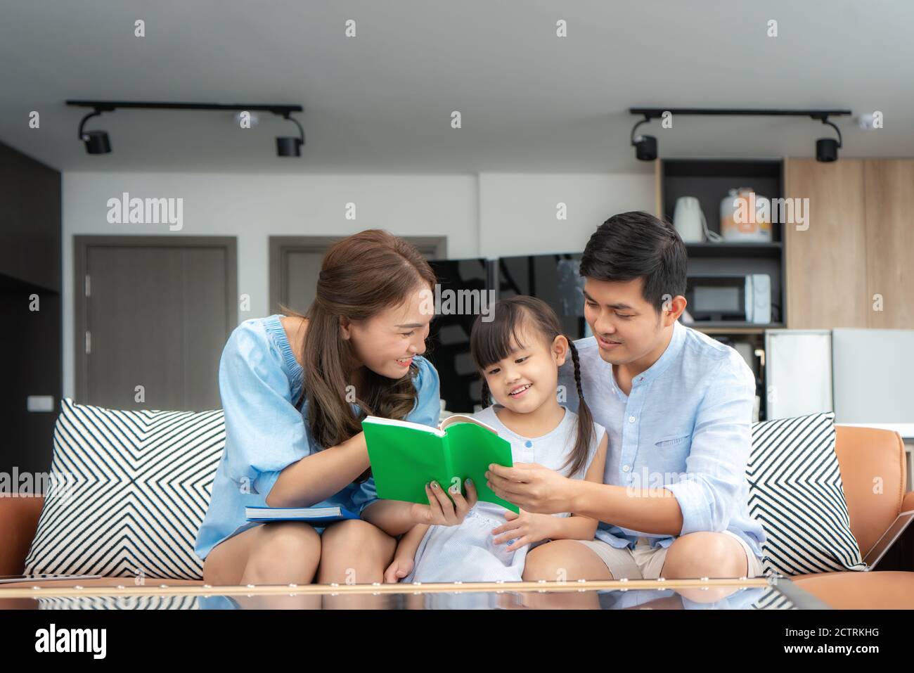 Famille asiatique avec son père, sa mère et sa fille lisant le livre d'histoire de queue de fées sur le canapé à la maison et pointant sur le livre le regardant avec happ Banque D'Images