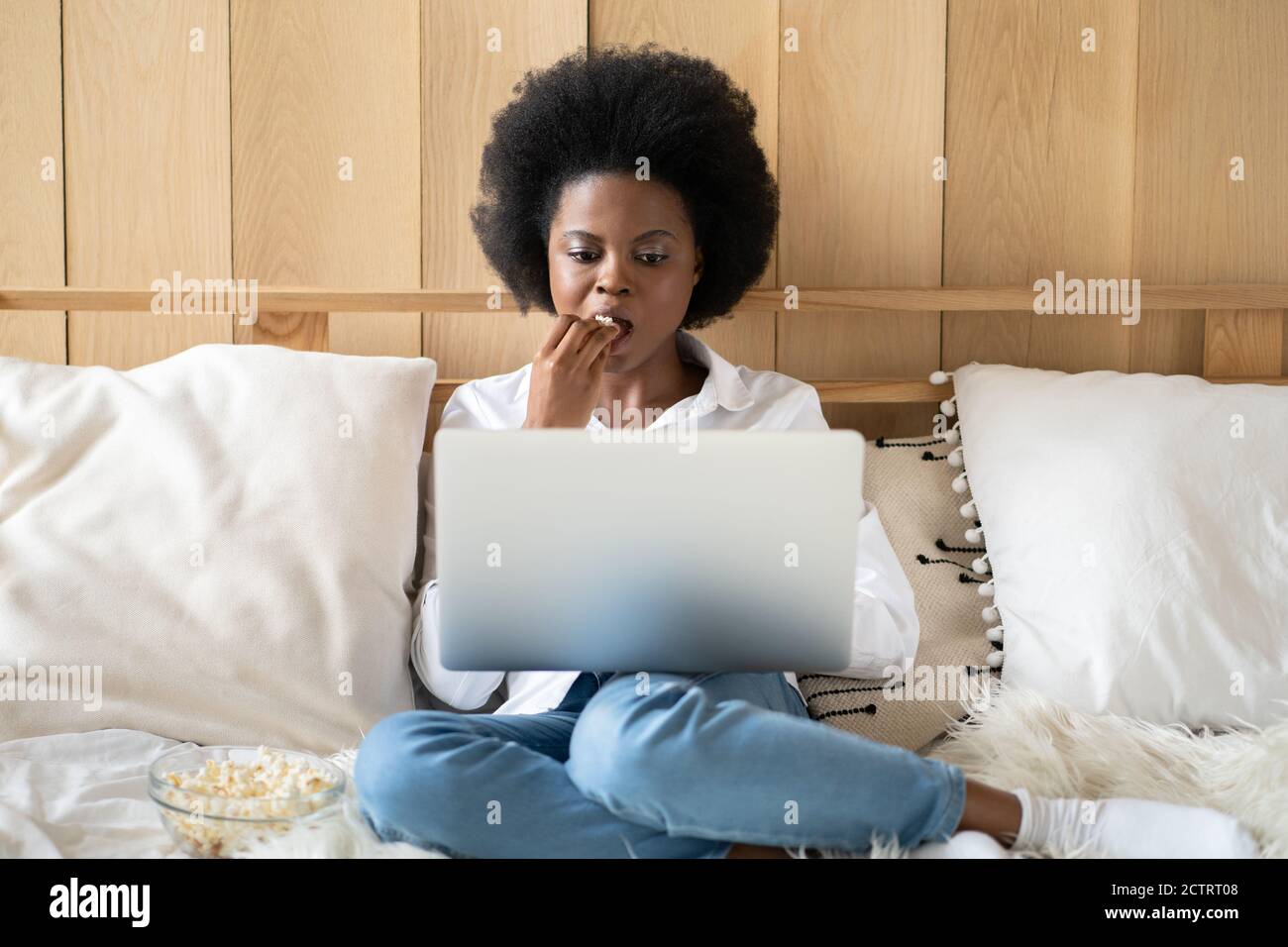 Femme afro-américaine concentrée en chemise blanche se détendant après le travail, mangeant du pop-corn tout en regardant un film d'horreur ou de thriller sur un ordinateur portable, allongé au lit. Paresseux r Banque D'Images