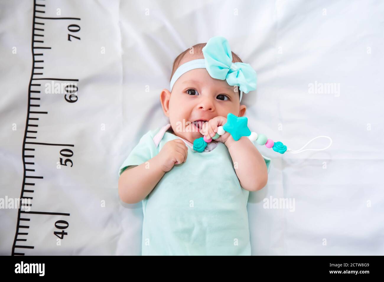 Un bébé de trois mois en vêtements vert menthe allongé sur un lit sur lequel est tiré une règle de mesure pour la croissance. Outil de dentition dans les mains, mastication Banque D'Images