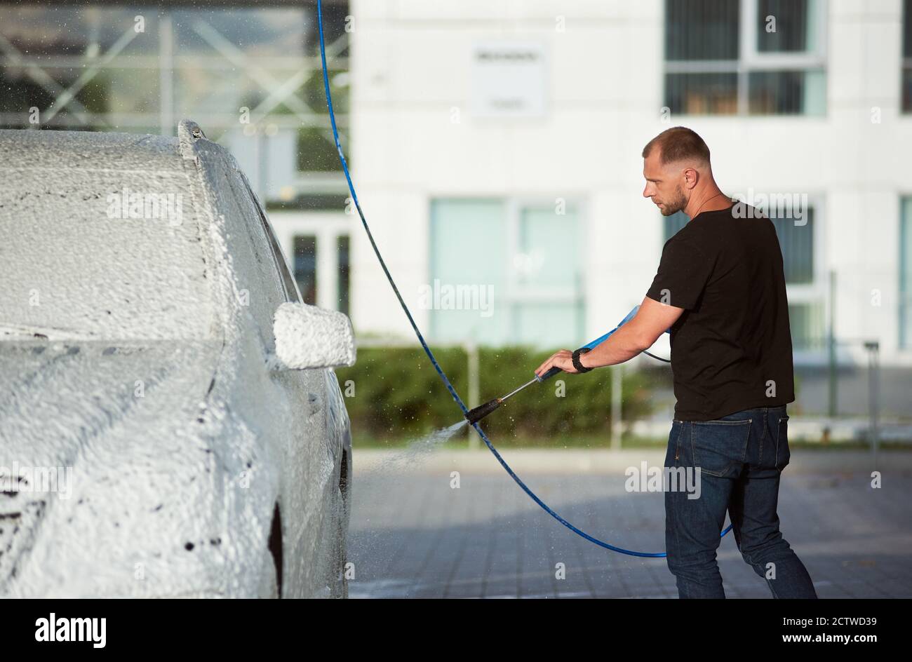 Un homme sérieux en t-shirts noirs lave sa voiture en tenant un tuyau avec de la mousse dehors, vue latérale Banque D'Images