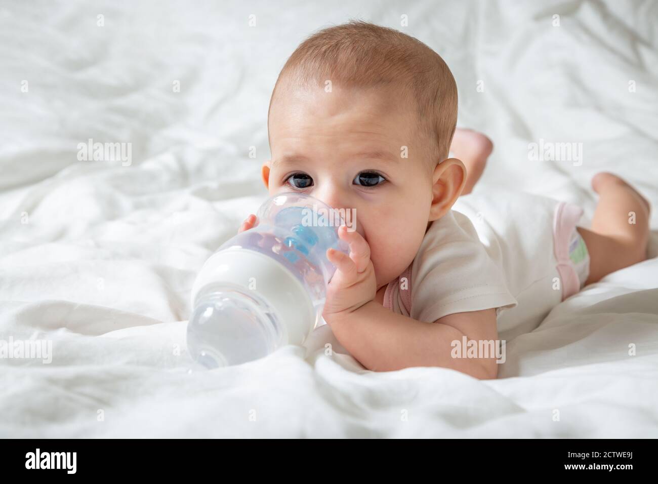 Bébé avec de grands yeux bruns allongé sur le lit blanc avec une bouteille d'eau spéciale avec mamelon. Essaie de la ronger Banque D'Images