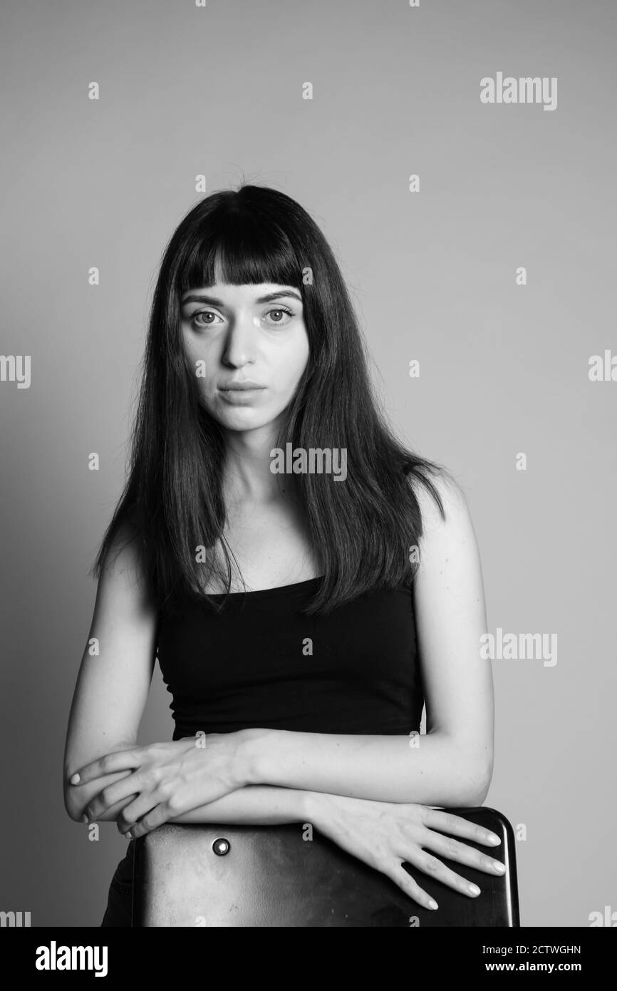 Studio portrait d'une jolie femme brune dans une veste noire, debout derrière la chaise, mains croisées, regardant sérieusement la caméra, contre une plaine Banque D'Images