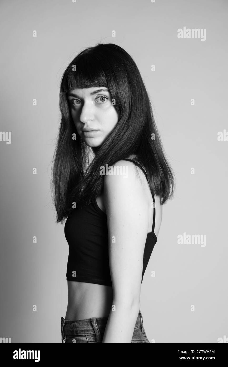 Portrait studio d'une jolie femme brunette dans un haut de bande spaghetti noir, regardant sérieusement l'appareil photo, debout à moitié tourné contre un gris Uni Banque D'Images