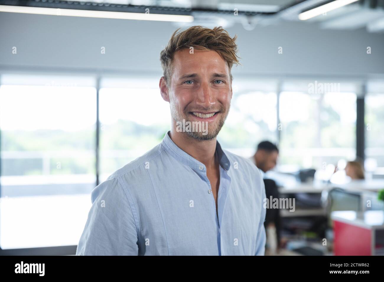 Portrait d'un homme d'affaires caucasien confiant avec des cheveux courts et une barbe souriante, avec des collègues travaillant en arrière-plan. Création d'entreprise Banque D'Images
