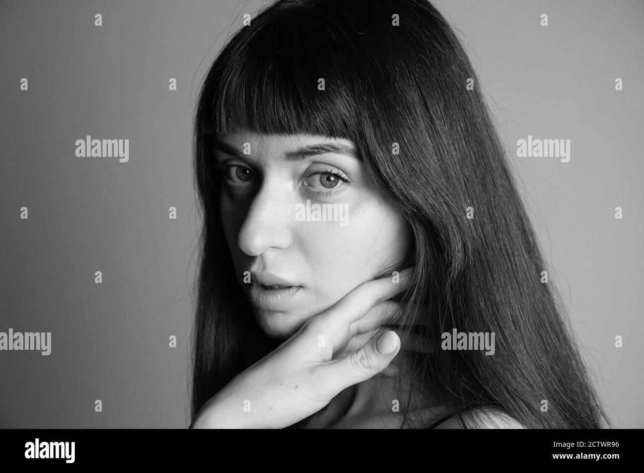 Gros plan portrait en studio d'une jolie femme brunette, main sur la joue, regardant l'appareil photo, à moitié tourné, sur un fond gris Uni Banque D'Images