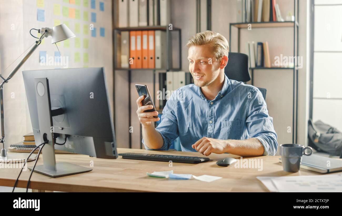 L'entrepreneur créatif assis à son bureau travaille sur un ordinateur de bureau dans le bureau élégant, choisit et commence à utiliser le smartphone, utilise l'application de médias sociaux Banque D'Images