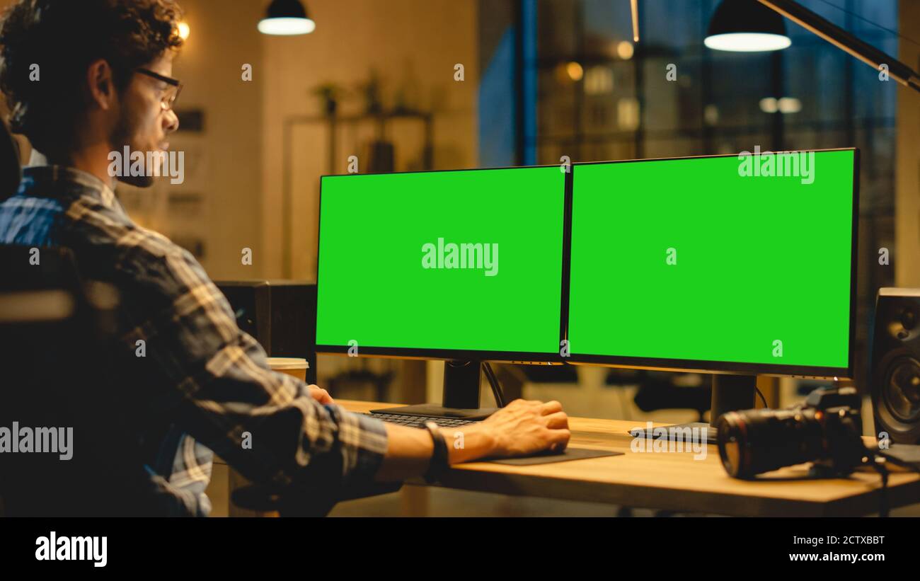 Au-dessus de l'épaule : le créateur assis à son bureau utilise un ordinateur de bureau avec deux écrans de maquette verts. Employé de bureau professionnel travaillant Banque D'Images