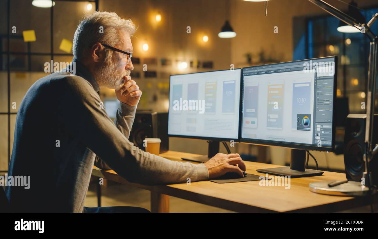 Créateur de logiciels mobiles d'âge moyen, assis à son bureau Utilise un ordinateur de bureau avec deux écrans affichant la conception d'application du smartphone Processus Banque D'Images