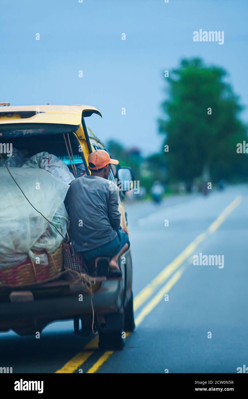 La vue arrière du porteur khmer est suspendue sur le minibus de livraison transporte une pleine charge de nourriture et de boissons sur la route rurale du Cambodge du Sud. Flou de mouvement. Banque D'Images