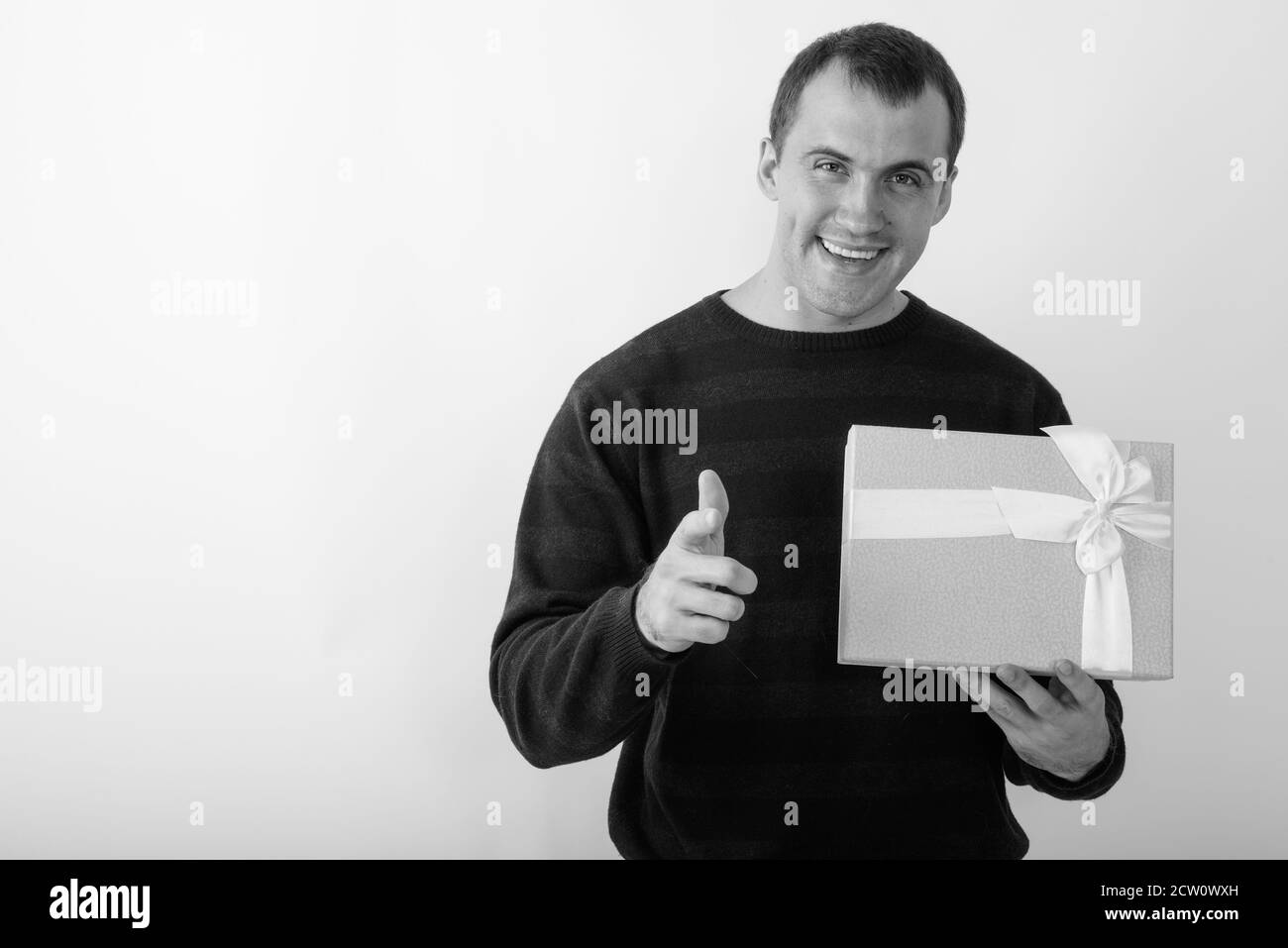 Portrait de jeune homme musclé tout en souriant heureux holding gift box and pointing at camera against white background Banque D'Images