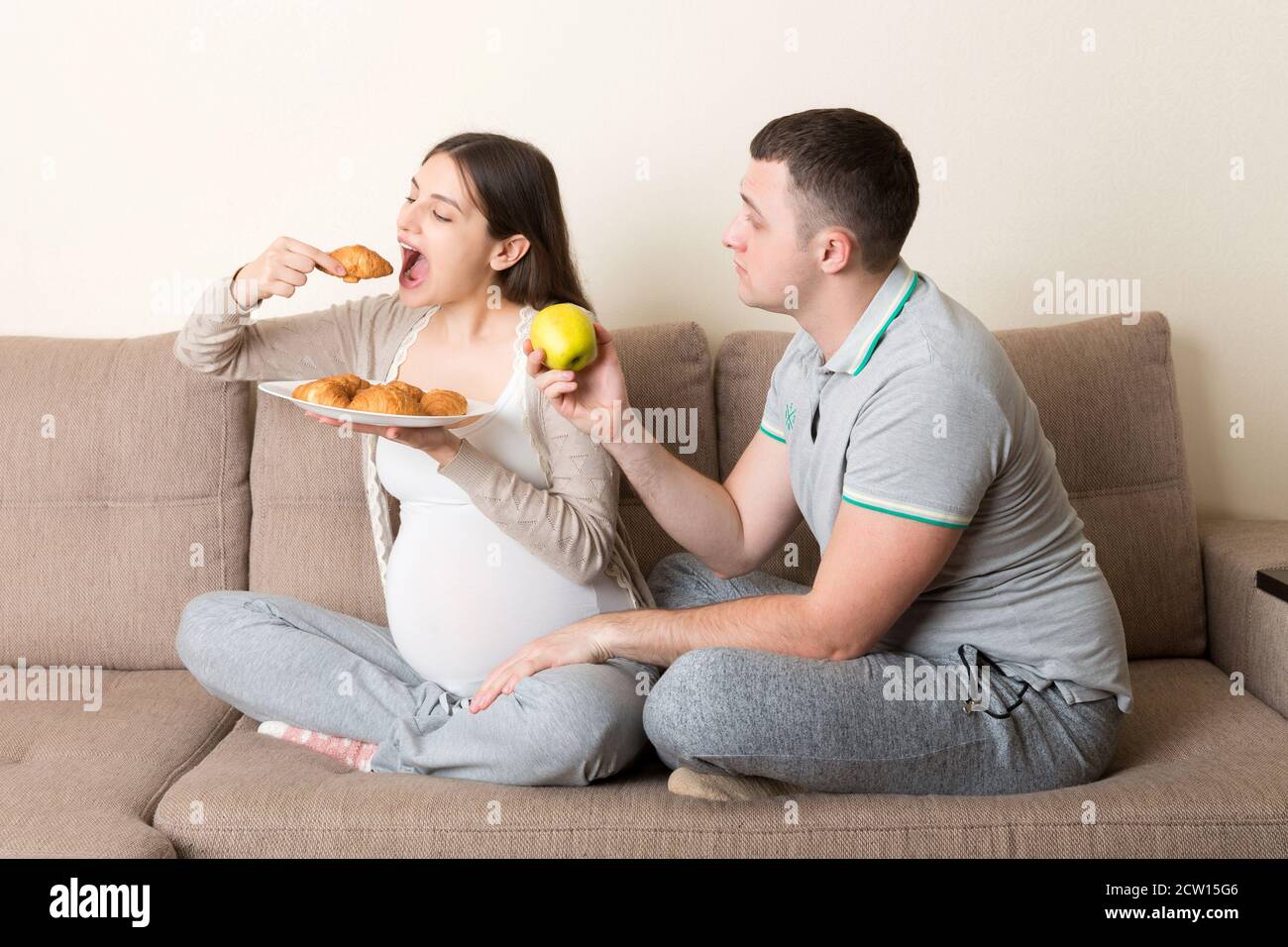 Le mari essaie de nourrir sa femme enceinte avec une pomme mais elle veut  un croissant assis sur le canapé à la maison. Régime alimentaire malsain  pendant la grossesse concept Photo Stock -
