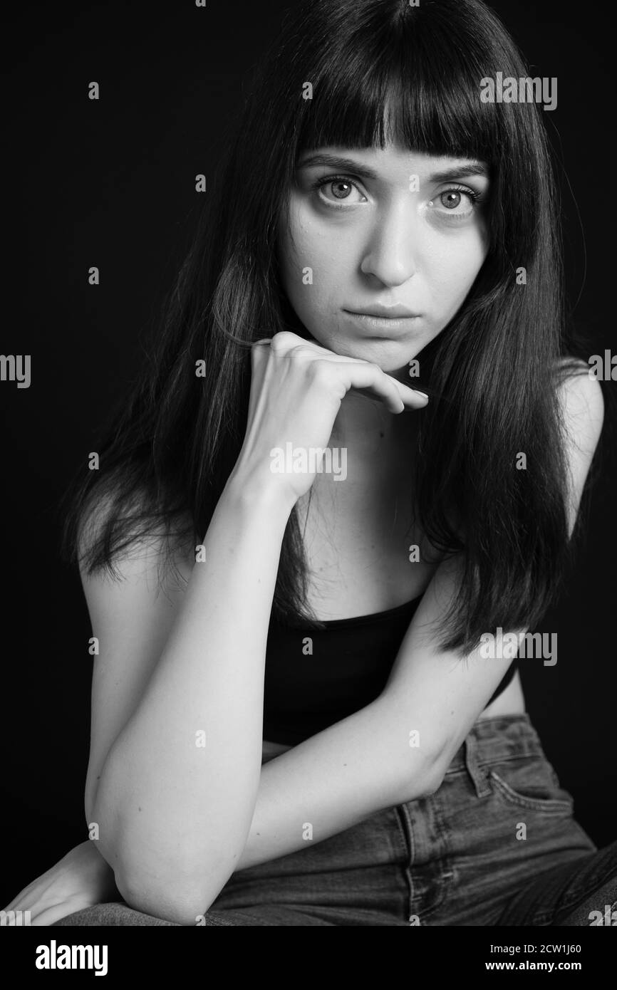 Portrait studio d'une jolie femme brune en haut à bandoulière spaghetti noir et jeans, regardant l'appareil photo, assis sur un fond noir Uni Banque D'Images