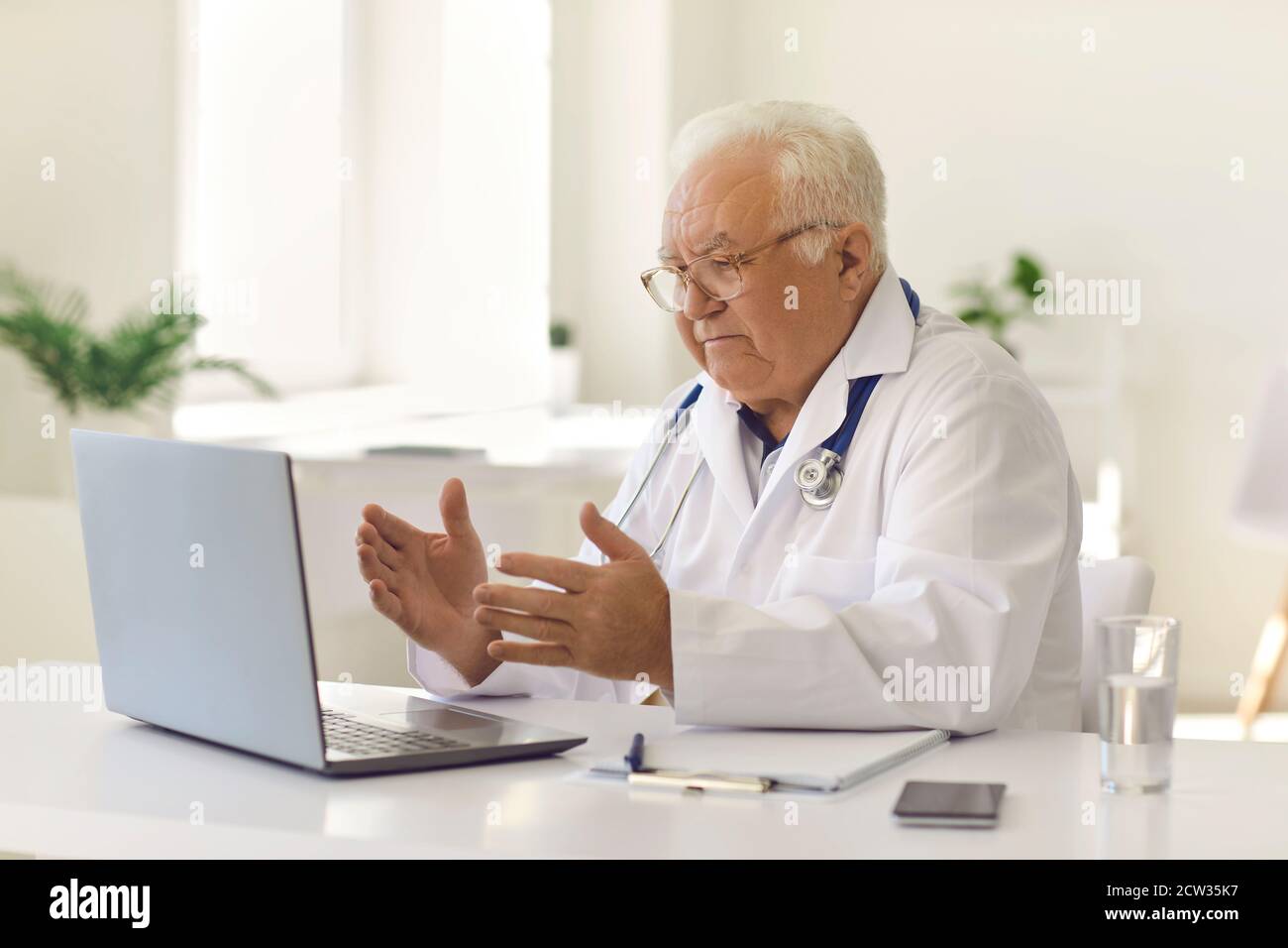 Médecin sénior sérieux assis à un bureau dans le bureau de l'hôpital et utiliser un ordinateur portable pour appeler ses patients par vidéo Banque D'Images