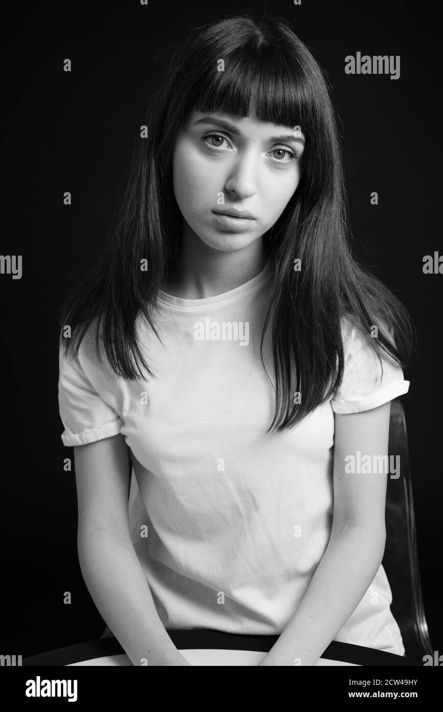 Portrait studio d'une jolie femme brune dans un t-shirt blanc, assis sur un fond noir Uni, regardant l'appareil photo Banque D'Images