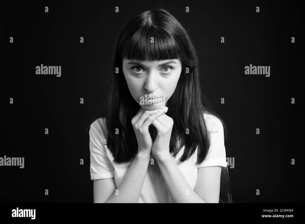 Portrait en studio d'une jolie femme brune dans un t-shirt blanc blanc, la tête reposant sur ses mains sur un fond noir Uni, regardant l'appareil photo Banque D'Images