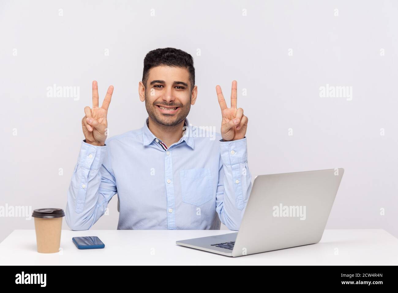 C'est gagnant ! Homme d'affaires joyeux assis au bureau avec un ordinateur portable sur le bureau, montrant le geste de victoire, célébrant le projet réussi, la promotion d'emploi. Banque D'Images