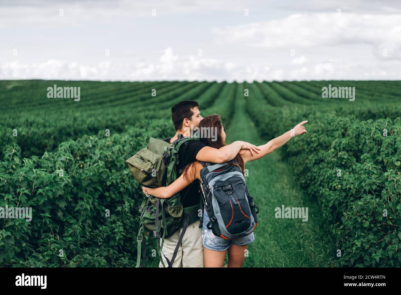 Vue arrière d'un jeune couple avec des sacs à dos sur les plantations de cassis. Une femme aux cheveux longs pointe sa main vers l'horizon. Banque D'Images