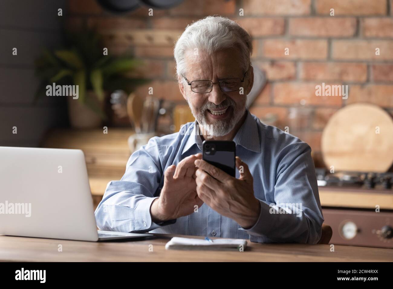 Un vieil homme souriant utilise un smartphone pour travailler sur un ordinateur portable Banque D'Images