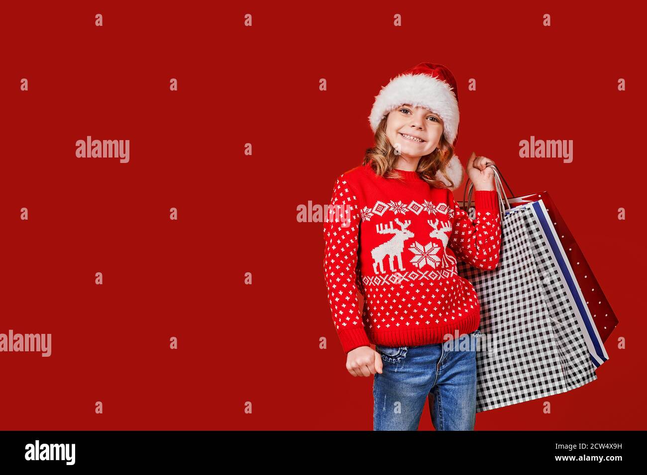 Adorable enfant en chapeau de père Noël rouge et chandail avec cerf portant des sacs de shopping colorés avec des cadeaux de Noël sur fond rouge. Banque D'Images
