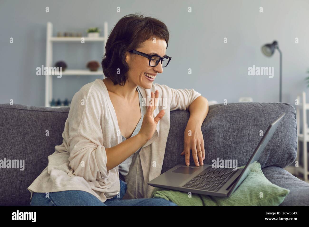 Femme souriante saluant quelqu'un pendant la conversation en ligne sur ordinateur portable à accueil Banque D'Images