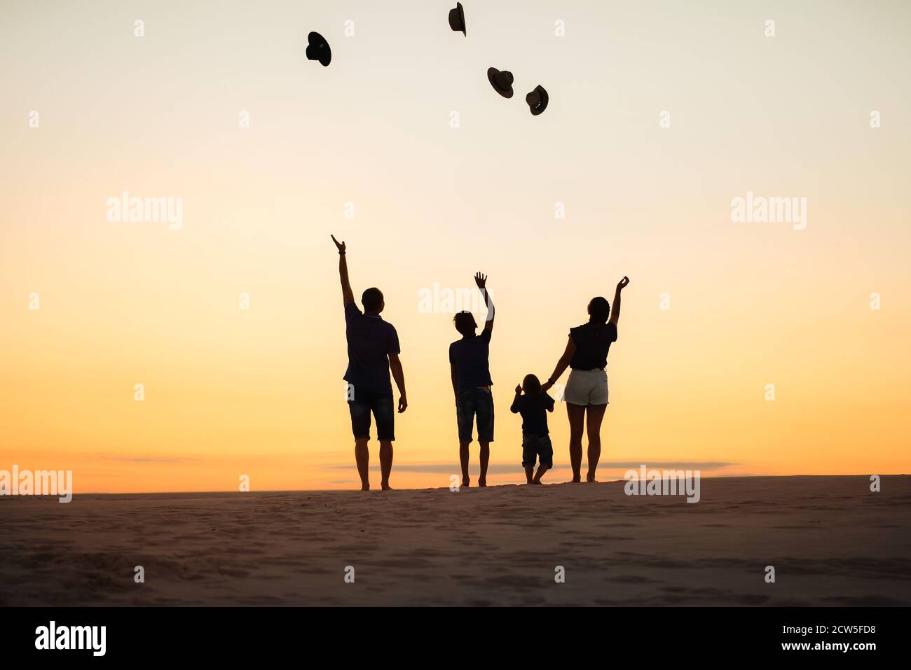 Silhouette de famille au coucher du soleil sur les sables. Maman, papa et deux fils jettent leurs chapeaux. Banque D'Images