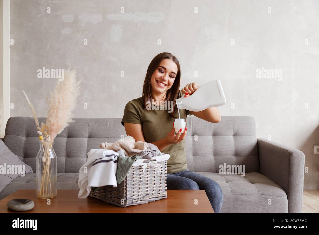 Femme versant du détergent dans le couvercle de la bouteille. Elle est assise à la table avec les vêtements recueillis dans le panier Banque D'Images