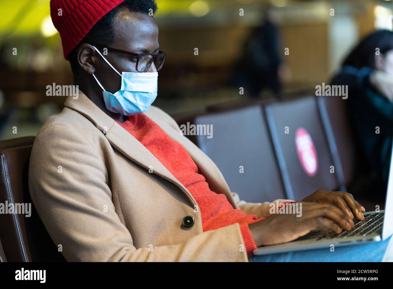 Élégant Noir voyageur homme millénaire porter un masque de protection du visage, assis dans le hall de l'aéroport, travaille à distance sur ordinateur portable tout en attendant un vol et b Banque D'Images